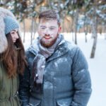 Couple standing in a snowy forest with snow on their faces and coats, illustrating the effects of winter weather on skin.