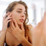 Young woman examining acne on her cheek in the mirror